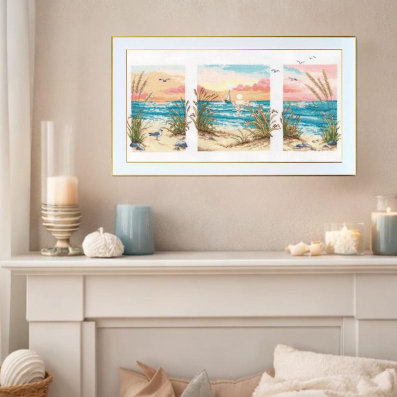 May include: A framed triptych cross-stitch artwork depicting a beach scene. The artwork features a sunset with a sailboat on the ocean, sand, and tall grass. The frame is white with a gold inner edge.