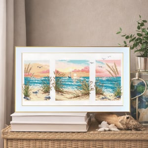 May include: A framed cross-stitch artwork depicting a beach scene with a sunset. The artwork is divided into three panels, showcasing a sailboat, seagulls, and beach grass. The color palette includes blues, pinks, and yellows, creating a serene coastal theme.