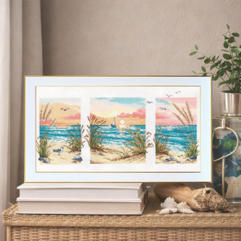 May include: A framed cross-stitch artwork depicting a beach scene with a sunset. The artwork is divided into three panels, showcasing a sailboat, seagulls, and beach grass. The color palette includes blues, pinks, and yellows, creating a serene coastal theme.