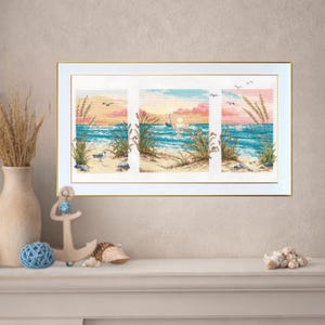 May include: A framed cross-stitch artwork depicting a beach scene with a sailboat at sunset. The artwork features a blue ocean, sandy shore, and tall grass. The frame is gold-colored, and the artwork is displayed on a white surface.