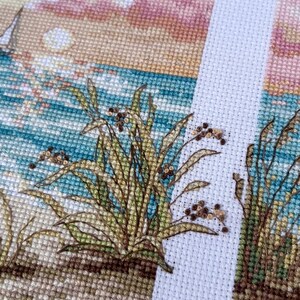 May include: Cross-stitch embroidery depicting a beach scene with a sailboat, ocean, and sunset. The foreground features detailed stitching of beach grass. The color palette includes blues, greens, tans, and pinks.