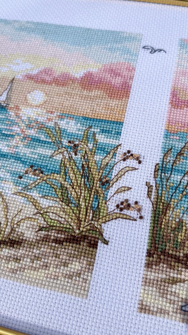 May include: Cross-stitch embroidery depicting a beach scene with a sailboat, ocean, and sunset. The foreground features detailed stitching of beach grass. The color palette includes blues, greens, tans, and pinks.