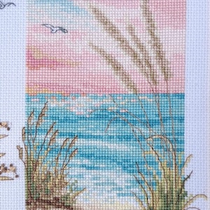 May include: Cross-stitch artwork depicting a serene beach scene. The sky transitions from pink to blue, with birds in flight. The ocean is turquoise, and the foreground shows sand, rocks, and tall grasses. A calming, nature-themed piece.