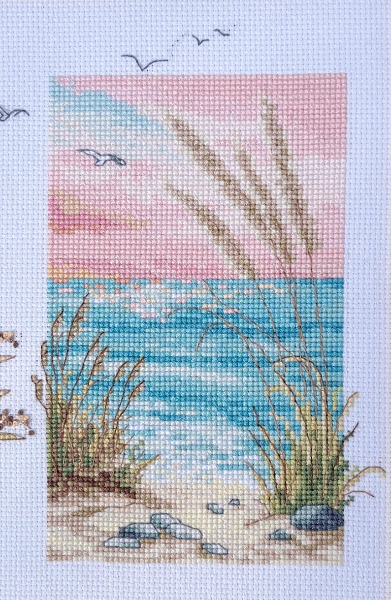 May include: Cross-stitch artwork depicting a serene beach scene. The sky transitions from pink to blue, with birds in flight. The ocean is turquoise, and the foreground shows sand, rocks, and tall grasses. A calming, nature-themed piece.