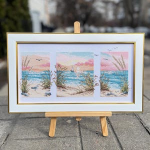 May include: A framed cross-stitch artwork depicting a beach scene with a sunset. The artwork features a white frame with a gold inner border. The scene includes a sailboat, birds, and beach grass. The artwork is displayed on a small wooden easel.