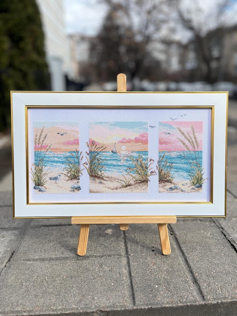 May include: A framed cross-stitch artwork depicting a beach scene with a sunset. The artwork features a white frame with a gold inner border. The scene includes a sailboat, birds, and beach grass. The artwork is displayed on a small wooden easel.