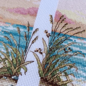 May include: Cross-stitch artwork depicting a coastal scene. The design features detailed stitching of beach grass, ocean waves, and a bird in flight against a pastel sky. The color palette includes shades of blue, beige, green, and pink.