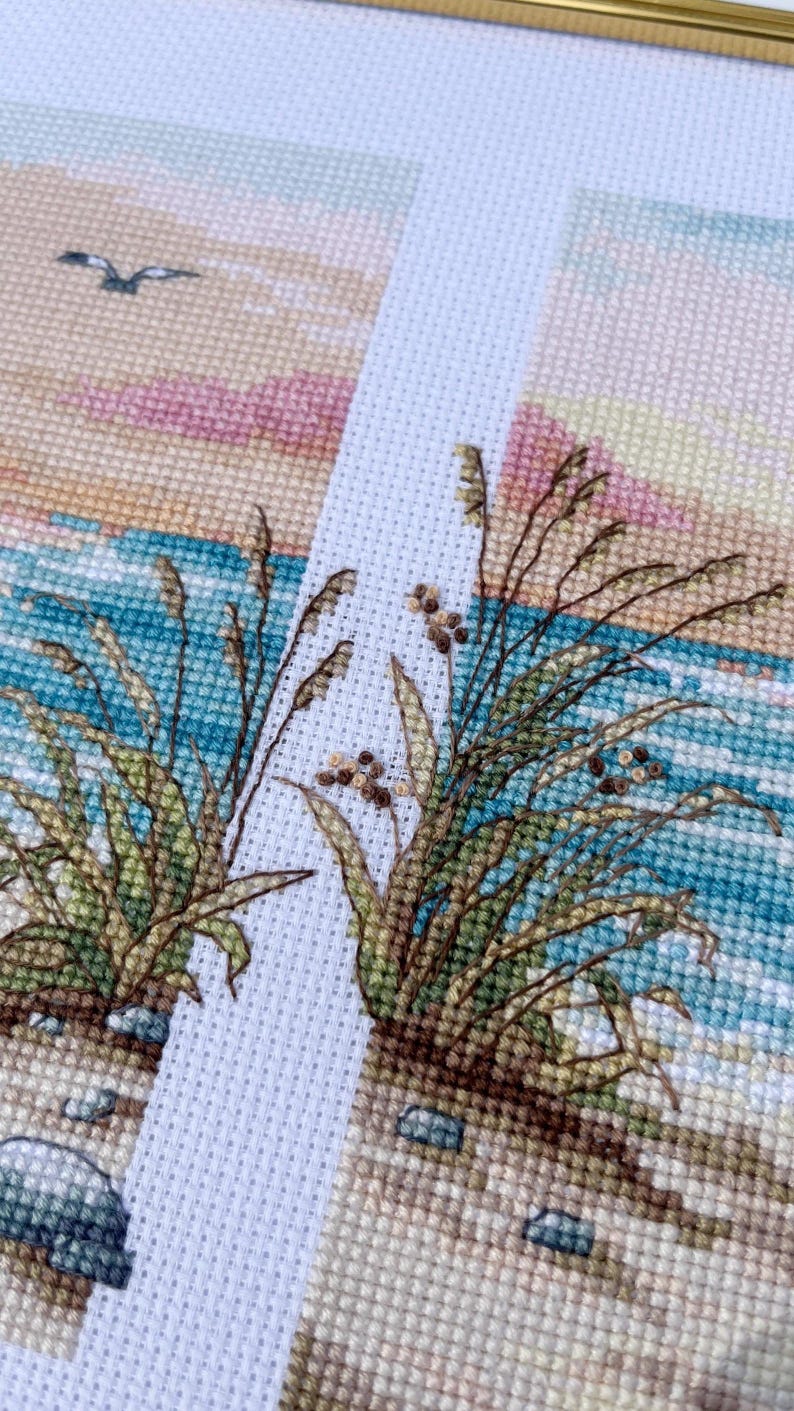 May include: Cross-stitch artwork depicting a coastal scene. The design features detailed stitching of beach grass, ocean waves, and a bird in flight against a pastel sky. The color palette includes shades of blue, beige, green, and pink.