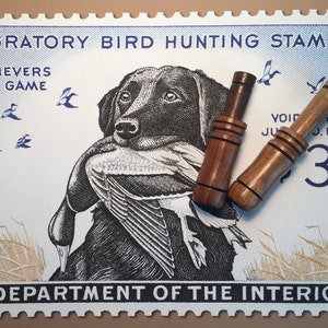 Pode incluir: Um selo vintage do Departamento do Interior dos Estados Unidos que representa um labrador retriever preto a recuperar um pato. O selo &eacute; azul e branco com o texto "Migratory Bird Hunting Stamp", "Retrievers Save Game", "Void After June 30, 1960", e "3".