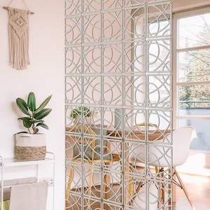 May include: A white room divider made of wood with a geometric pattern. The divider is made up of individual squares with a circular design. The divider is standing in front of a window with a view of a city.
