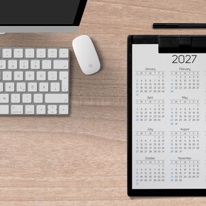 May include: A black clipboard holding a 2027 calendar with twelve months displayed in a grid. The calendar features black text on a white background, showing the year 2027 at the top, followed by monthly grids from January through December.