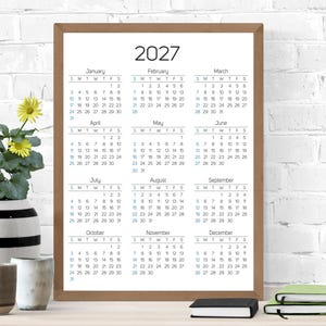 May include: A vertical wall calendar for the year 2027 displayed in a simple brown wooden frame. The calendar features a white background with black text for the months and days of the week, and blue text for specific dates, organized in a grid.
