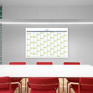 May include: A large white 2027 calendar with blue and green accents hangs on a grey wall. Below, a white table is surrounded by red chairs. The room has a modern, minimalist design with a glass block wall.