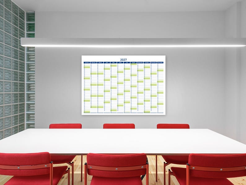 May include: A large white 2027 calendar with blue and green accents hangs on a grey wall. Below, a white table is surrounded by red chairs. The room has a modern, minimalist design with a glass block wall.