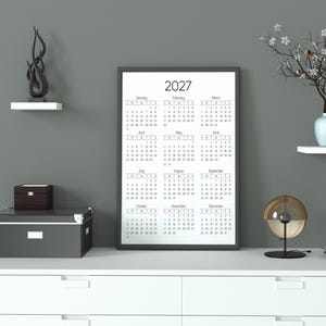 May include: A vertical wall calendar for the year 2027 with a black frame, displaying all twelve months in a grid layout. The calendar features black text on a white background, showing the months from January to December with days of the week.