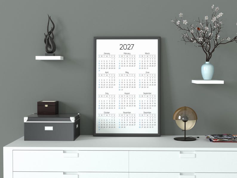 May include: A vertical wall calendar for the year 2027 with a black frame, displaying all twelve months in a grid layout. The calendar features black text on a white background, showing the months from January to December with days of the week.