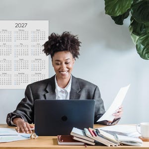 May include: A wall calendar for the year 2027 featuring a grid of twelve months from January to December. The calendar has a white background with black text for the months and days, and blue text for Sundays. It is displayed on a wall.