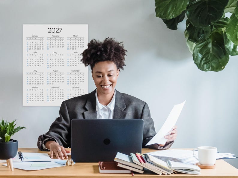 May include: A wall calendar for the year 2027 featuring a grid of twelve months from January to December. The calendar has a white background with black text for the months and days, and blue text for Sundays. It is displayed on a wall.
