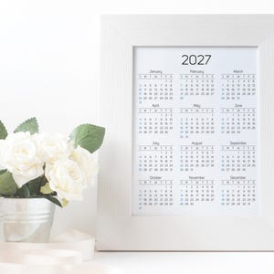 May include: A white picture frame containing a 2027 calendar with twelve months displayed in a grid format. Next to the frame is a small silver metal bucket filled with white artificial roses and green leaves, with a white ribbon draped nearby.