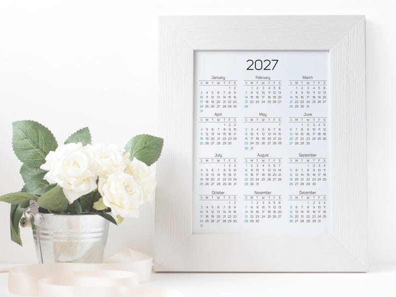 May include: A white picture frame containing a 2027 calendar with twelve months displayed in a grid format. Next to the frame is a small silver metal bucket filled with white artificial roses and green leaves, with a white ribbon draped nearby.