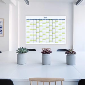 May include: A large 2027 year planner with a blue and green grid hangs on a white wall above a white table. Three small grey pots with succulents sit on the table. An abstract framed art piece is on the wall.