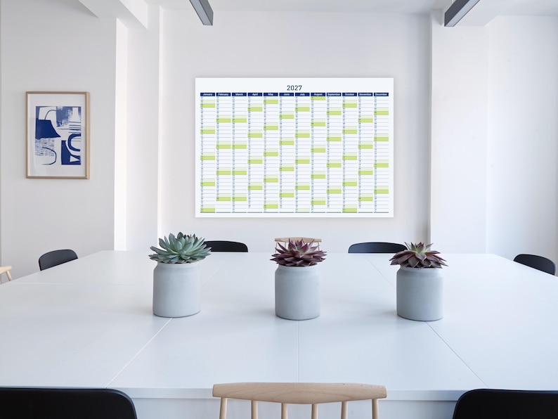 May include: A large 2027 year planner with a blue and green grid hangs on a white wall above a white table. Three small grey pots with succulents sit on the table. An abstract framed art piece is on the wall.