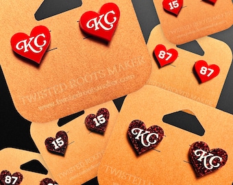 May include: Red heart-shaped earrings with white text, including "KC" and numbers "15" and "87", displayed on tan cardboard earring cards. The cards have a hole for hanging and the text "TWISTED ROOTS MAKER".