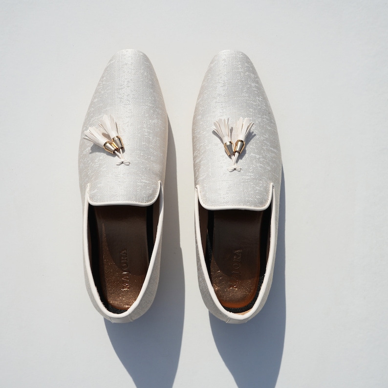 May include: A pair of white satin loafers with gold tassels. The loafers have a brown leather insole and are made by the brand "Marotta".