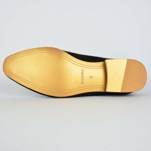 May include: The bottom of a black suede shoe with a gold sole. The sole has the brand name "MAJORA" and the size "8" printed on it.