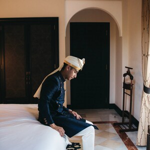 May include: A man wearing a white turban with gold trim sits on a white bed, wearing a dark blue long-sleeved shirt with gold buttons and white pants. He is wearing white loafers with gold accents.