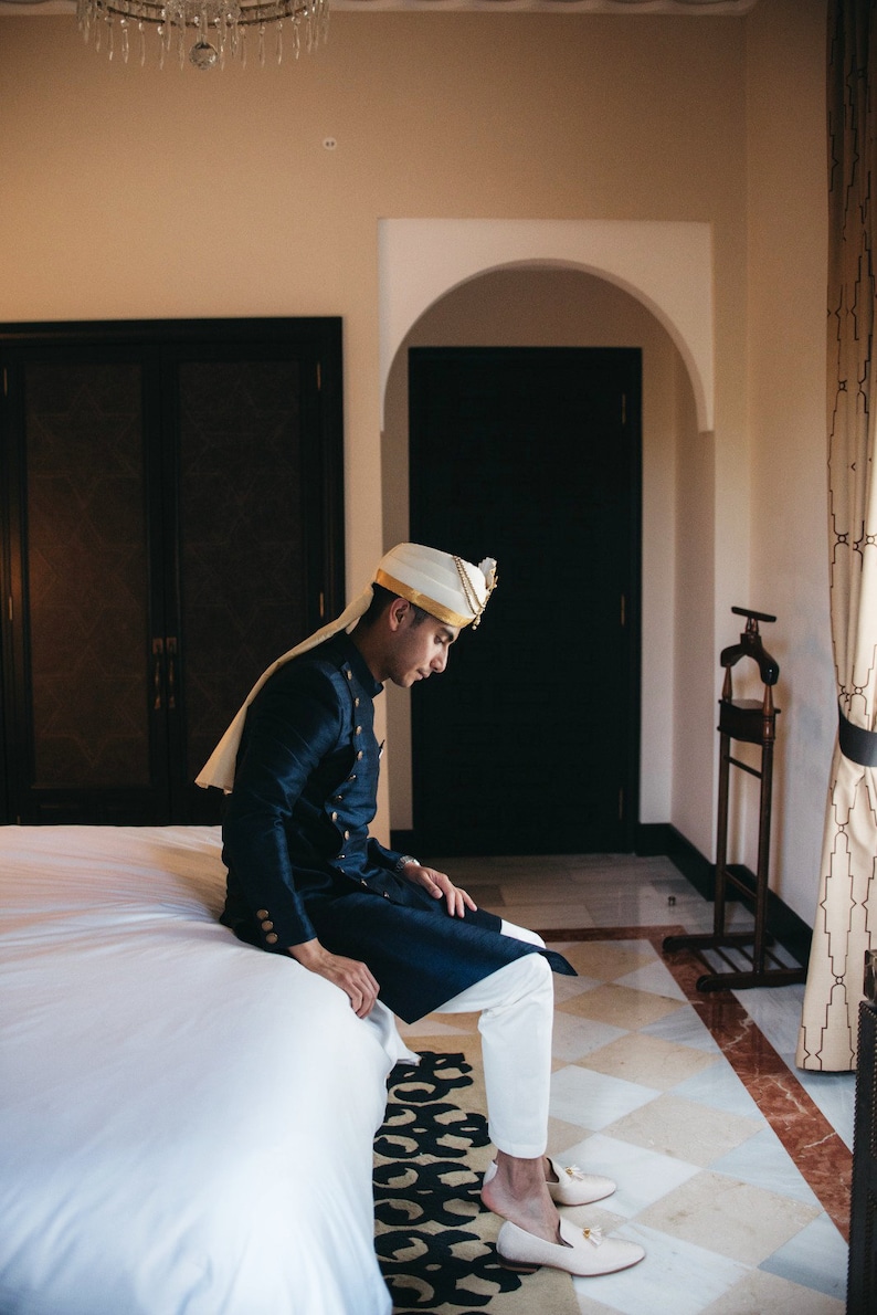May include: A man wearing a white turban with gold trim sits on a white bed, wearing a dark blue long-sleeved shirt with gold buttons and white pants. He is wearing white loafers with gold accents.