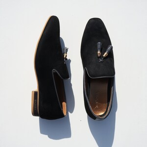 May include: A pair of black suede loafers with gold tassel accents. The loafers have a brown leather sole and the brand name "MAJORA" is printed on the side.
