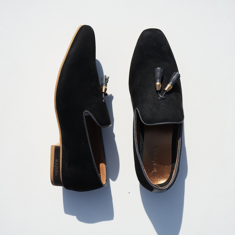 May include: A pair of black suede loafers with gold tassel accents. The loafers have a brown leather sole and the brand name "MAJORA" is printed on the side.