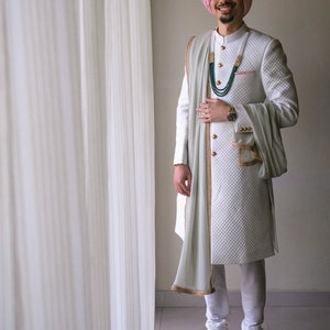 May include: A man wearing a white quilted sherwani with gold trim, a light green scarf, and a pink turban. He is wearing a green and gold beaded necklace and a watch on his left wrist.