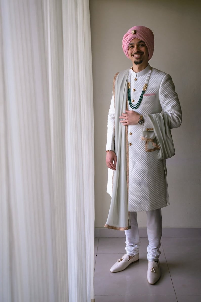 May include: A man wearing a white quilted sherwani with gold trim, a light green scarf, and a pink turban. He is wearing a green and gold beaded necklace and a watch on his left wrist.