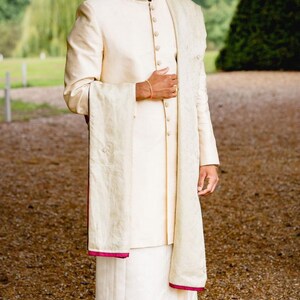 May include: A man wearing a white traditional Indian outfit with a gold turban and a white shawl. He is standing in a garden setting.