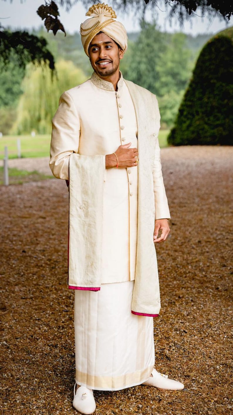 May include: A man wearing a white traditional Indian outfit with a gold turban and a white shawl. He is standing in a garden setting.