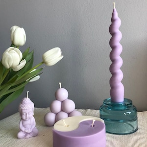 May include: A set of lavender scented candles in various shapes, including a meditating Buddha, a yin yang design, a cluster of spheres, and a tall, twisted candle. The candles are displayed on a white surface with a bouquet of white tulips in the background.