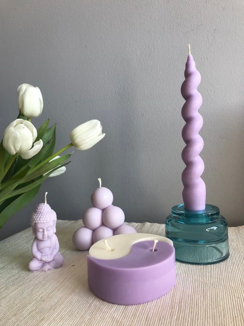 May include: A set of lavender scented candles in various shapes, including a meditating Buddha, a yin yang design, a cluster of spheres, and a tall, twisted candle. The candles are displayed on a white surface with a bouquet of white tulips in the background.