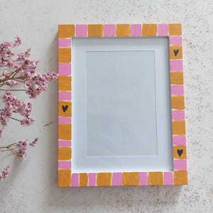 Op de afbeelding: Een rechthoekige fotolijst met een roze en gouden geblokte rand en zwarte hartaccenten. De lijst heeft een witte binnenkant en staat tegen een getextureerde witte achtergrond. Gedroogde roze bloemen bevinden zich in de linkerbovenhoek.