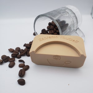 May include: A tan-colored silicone mold for making espresso cups. The mold has the words "Espresso Yourself" embossed on the top and a stylized coffee cup design on the bottom. Coffee beans are scattered around the mold.