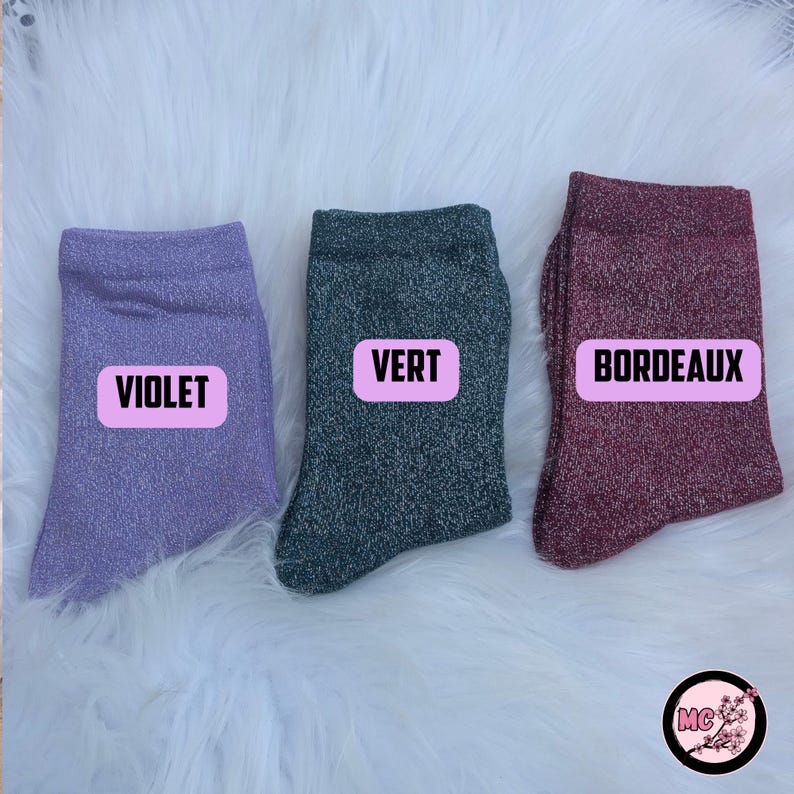 May include: Three pairs of sparkly socks in violet, green, and burgundy. Each sock has a rectangular label with the color name in white text. The socks are displayed on a white, fluffy surface.