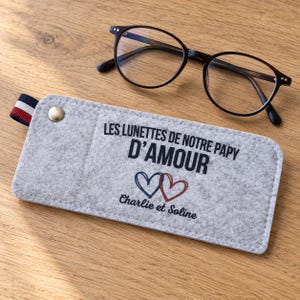 Könnte beinhalten: Ein hellgraues Brillenetui aus Filz mit dem Text LES LUNETTES DE NOTRE PAPY D'AMOUR, zwei Herzumrissen in Blau und Rot sowie den Namen Charlie et Soline, dazu eine Brille mit schwarzem Rahmen und einer gestreiften Stofflasche.
