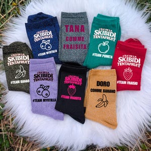 May include: A collection of colorful socks with various designs and text. Each sock features the words "L'ILE DE LA SKIBIDI TENTAFRUIT" and a different fruit graphic, such as a banana, apple, or strawberry. The socks are arranged on a fluffy white surface.