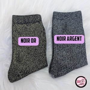 May include: Two pairs of black socks with metallic threads. One pair is labeled "NOIR OR" in a pink rectangle, the other is labeled "NOIR ARGENT". The socks are displayed on a white, fluffy surface.