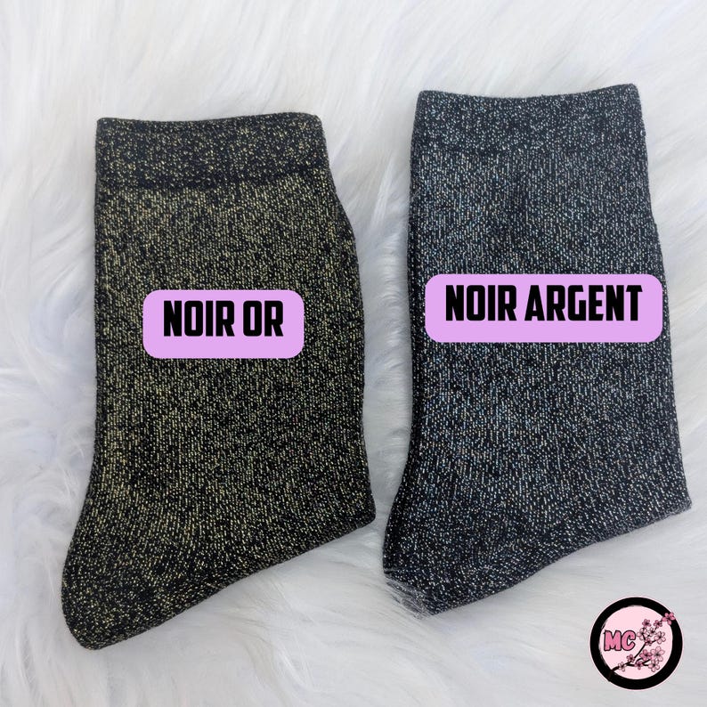 May include: Two pairs of black socks with metallic threads. One pair is labeled "NOIR OR" in a pink rectangle, the other is labeled "NOIR ARGENT". The socks are displayed on a white, fluffy surface.