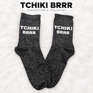 Pode incluir: Um par de meias pretas com glitter apresentando o texto branco TCHIKI BRRR impresso na parte superior. As meias possuem um punho canelado no topo e um acabamento cintilante e brilhante por todo o tecido, apresentando uma textura distinta.