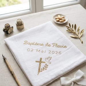 Könnte beinhalten: Weißes Tauf-Taschentuch aus Leinen mit goldener Stickerei. Der Text lautet "Baptême de Flavie" und "02 Mai 2026". Ein Kreuz, eine Taube und ein Olivenzweig sind ebenfalls gestickt. Tintenfässer, ein Stift und dekorative Elemente sind darum angeordnet.