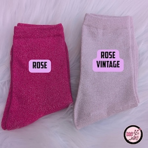May include: Two pairs of socks, one hot pink and one light pink, with the text "ROSE" and "ROSE VINTAGE" on labels. The socks are made of a shiny material and are displayed on a white, fluffy surface.