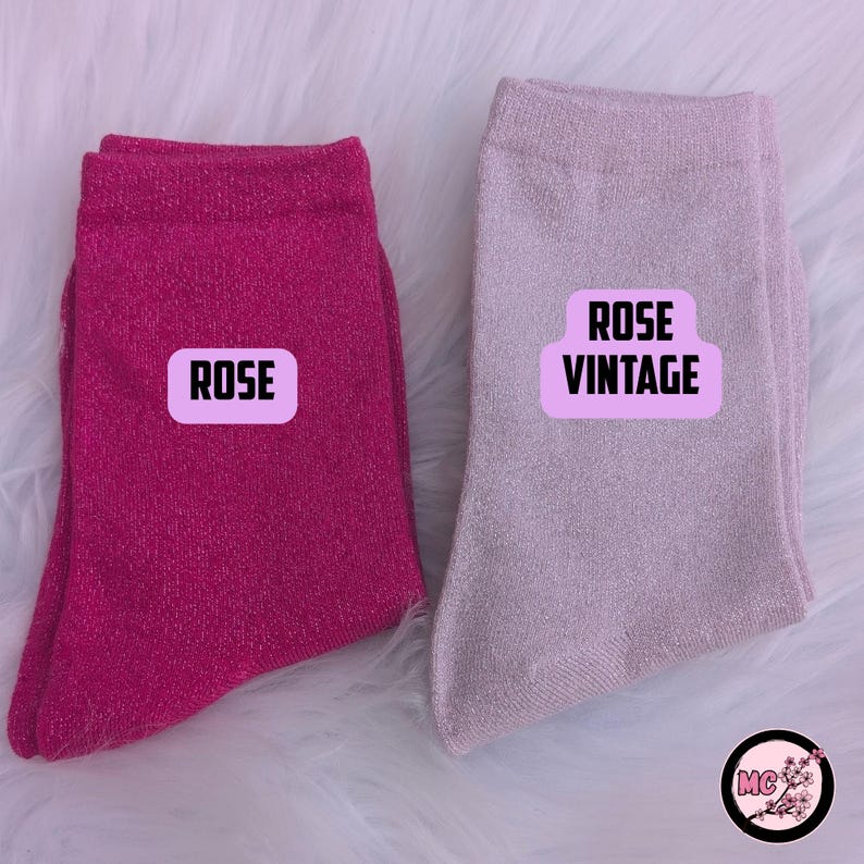 May include: Two pairs of socks, one hot pink and one light pink, with the text "ROSE" and "ROSE VINTAGE" on labels. The socks are made of a shiny material and are displayed on a white, fluffy surface.